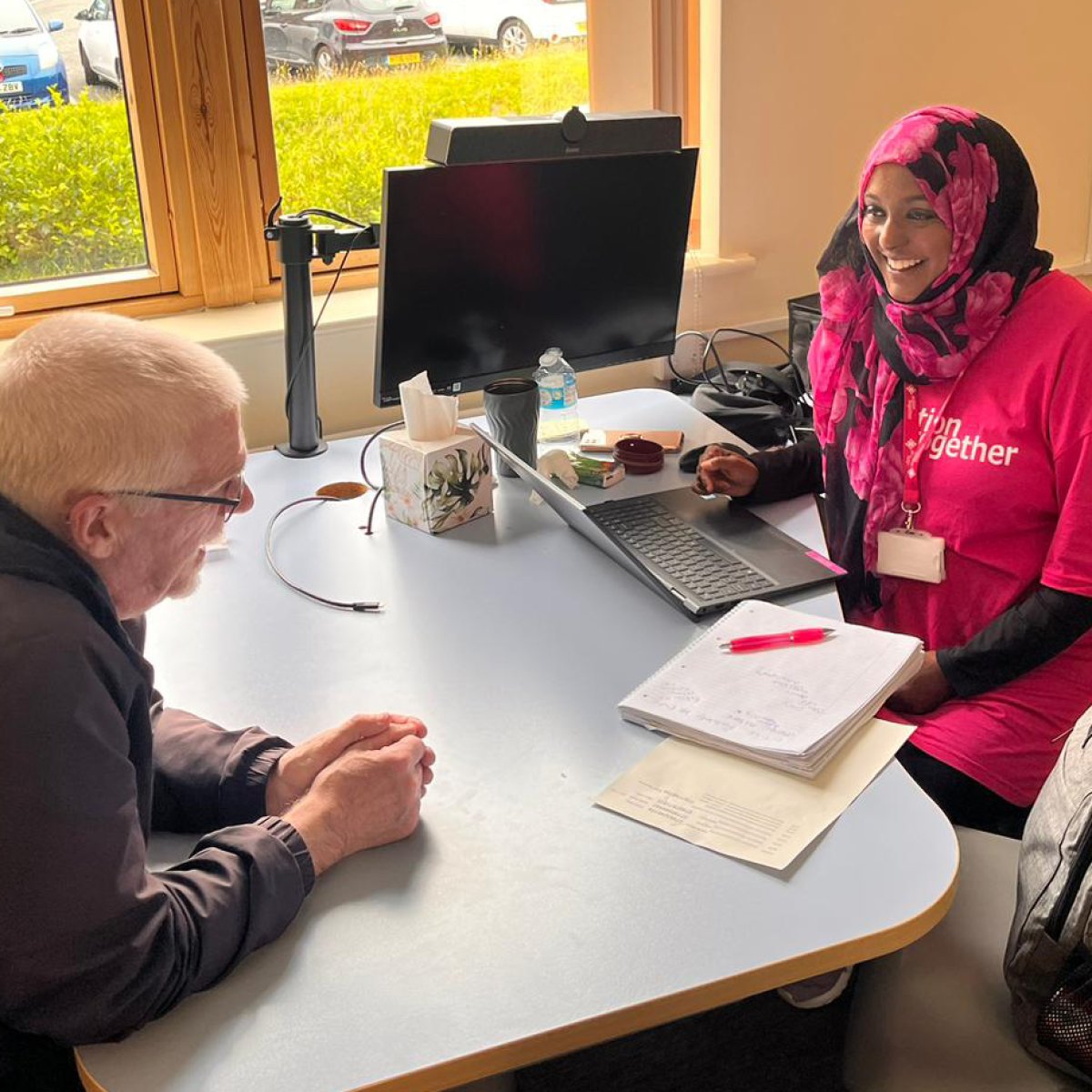 Woman (Asma) in pink, Action Together t-shirt chatting with man at table