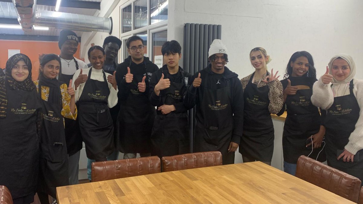 Group of young students in black cooking aprons smiling at camera with thumbs up