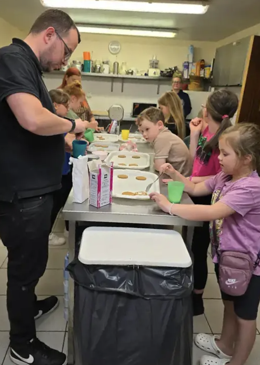 Group of children in industrial kitchen carrying out baking activities at a table, guided by three adult helpers