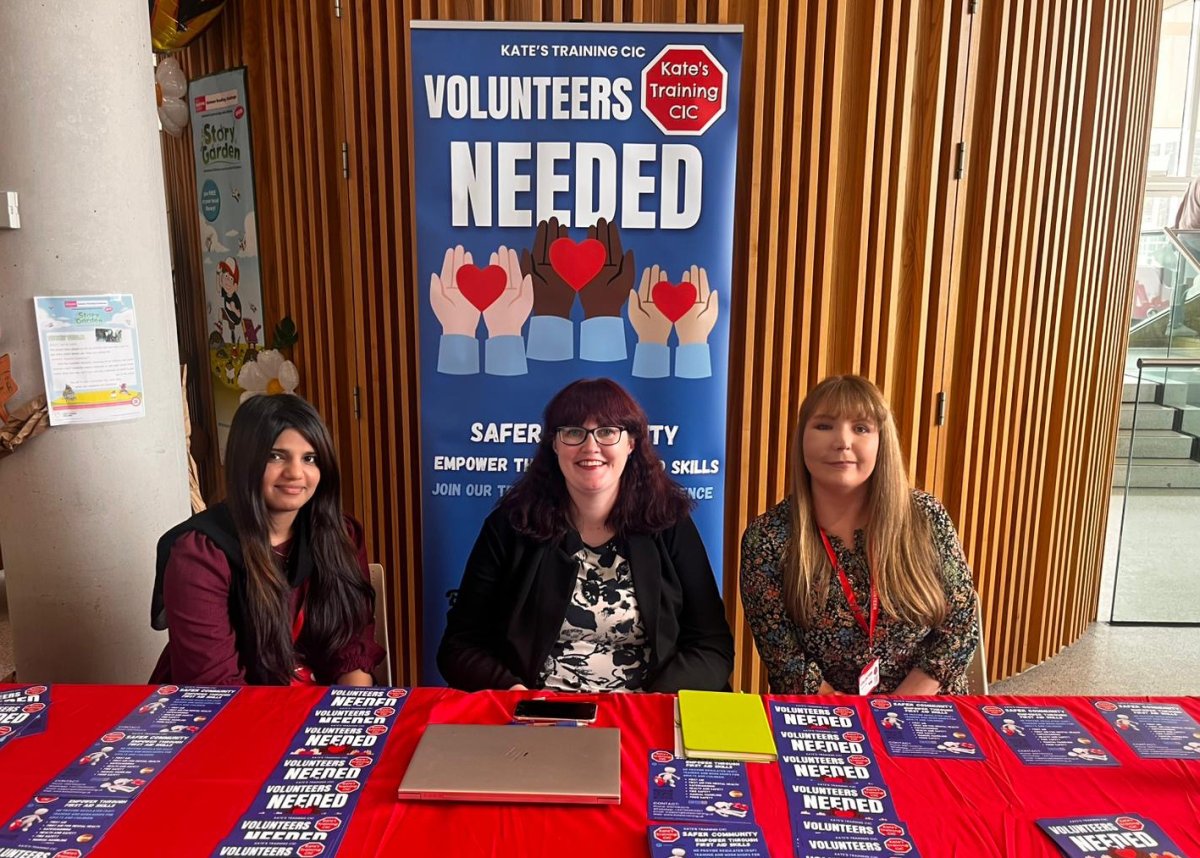 Three women sat behind exhibition table and banner advertising 'volunteers needed' for Kates Training CIC in Rochdale